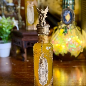 May include: A decorative glass bottle filled with yellow liquid, labeled "Pixie Dust Designs." The bottle has a gold-colored fairy figurine on the stopper and is adorned with gold accents. The bottle is approximately 8 inches tall.