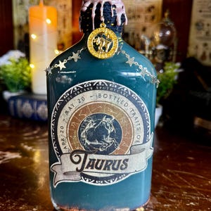 May include: A teal-colored apothecary bottle with a decorative label featuring the Taurus zodiac symbol. The bottle is adorned with a gold charm and star-shaped chain. The bottle's neck is sealed with a pink wax.