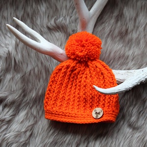 May include: Orange crocheted beanie with a pom pom on top and a wooden deer charm attached to the side.