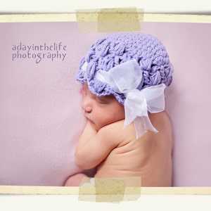 Puede incluir: Un bebé durmiendo con un gorro de crochet morado con una cinta blanca. El bebé está acostado sobre una manta de color morado claro.