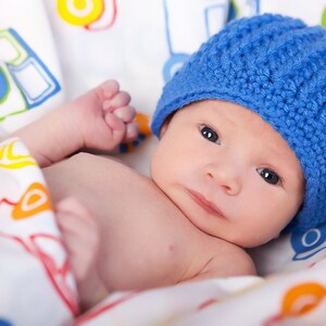 May include: A newborn baby wearing a blue knitted hat lies on a white blanket with a colorful car pattern.