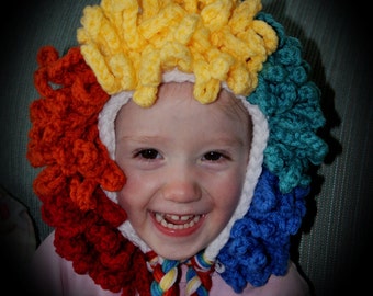 May include: A child wearing a colorful crocheted hat with red, yellow, blue, and green yarn. The hat is shaped like a clown's hair. The child is smiling.