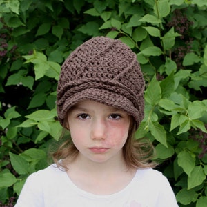 May include: A young person wearing a brown crocheted hat with a brim. The hat has a braided design and is worn over the person's brown hair. The person is wearing a white shirt and has a neutral expression.