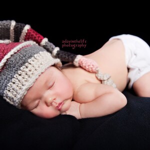 Op de afbeelding: Een baby die een veelkleurige gehaakte muts draagt met een roze en witte gehaakte bloem aan de zijkant, slaapt op een zwart oppervlak.