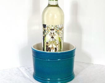 Könnte beinhalten: Eine Flasche Weißwein mit grünem Verschluss steht in einem türkisfarbenen Weinkühler aus Keramik. Der Kühler ist zylindrisch mit hellbeigem Innenraum und glänzendem Finish. Die Weinflasche hat ein dekoratives Etikett.