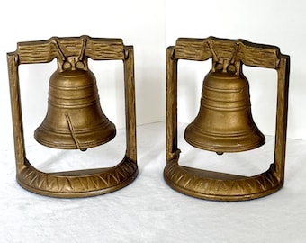 May include: Two bronze-colored bookends, each featuring a bell design within a rectangular frame. The bells have a detailed, textured surface and are suspended from a wooden-like top. The base of each bookend has a decorative, repeating pattern.