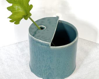 Op de afbeelding: Een turquoise keramische vaas met een uniek ontwerp, met een cilindrisch lichaam en een gedeeltelijk bedekte bovenkant. Een groen blad is in de vaas geplaatst. De vaas heeft een getextureerd oppervlak en een kleine, afgeronde basis.
