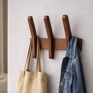 May include: A wooden wall-mounted coat rack with three hooks. A beige tote bag hangs from one hook, and a blue denim jacket hangs from another. The rack is mounted on a light gray wall.