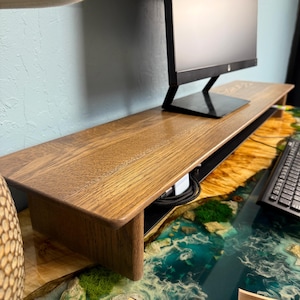 May include: A wooden monitor stand with a black cable management system. The stand is sitting on a blue and green resin table with a white and brown pattern.