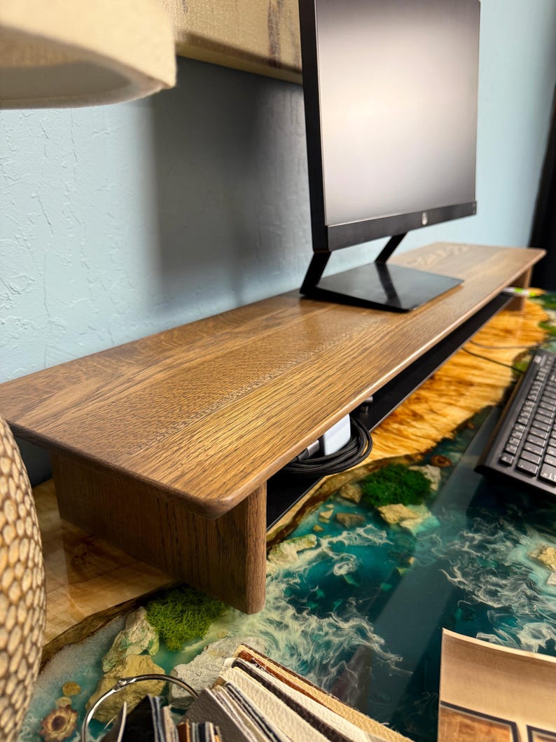May include: A wooden monitor stand with a black cable management system. The stand is sitting on a blue and green resin table with a white and brown pattern.