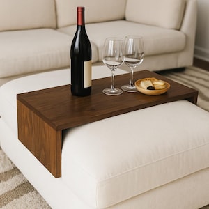 Op de afbeelding: Een donkerhouten ottomaantray met een fles rode wijn, twee wijnglazen en een kleine houten kom met snacks. De tray staat op een witte ottoman, met een beige bank op de achtergrond.