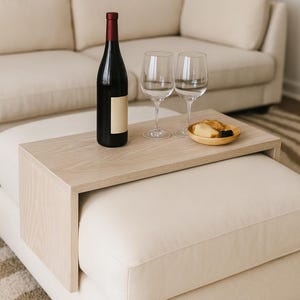 Peut inclure: Une bouteille de vin noire avec une étiquette vierge et un bouchon rouge, deux verres à vin transparents et une petite assiette en bois avec de la nourriture sont sur un plateau en bois clair. Le plateau est posé sur un pouf blanc, avec un canapé beige en arrière-plan.