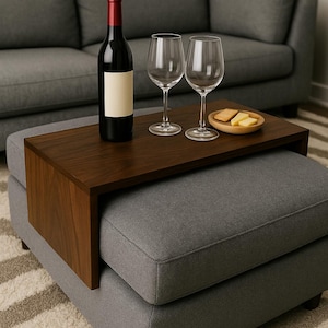 May include: A dark wood ottoman tray holds a bottle of red wine, two wine glasses, and a small wooden dish with cheese. The tray sits on a grey upholstered ottoman, with a grey sofa in the background. The scene suggests a relaxing evening.