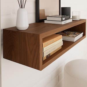 Puede incluir: Estantería flotante rectangular de madera marrón oscuro. La estantería contiene libros, una vela negra, un jarrón blanco y un pequeño espejo. La estantería está montada en una pared de ladrillo blanco.