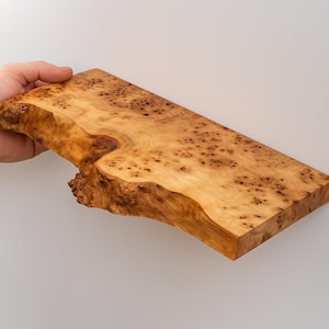 May include: A rectangular, unfinished wooden slab with a natural edge. The wood has a light brown color with darker brown spots and a unique grain pattern. The slab is being held by a hand.