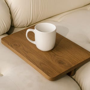 May include: A white ceramic mug with vertical ribbed detailing sits on a rectangular wooden tray. The tray is a warm brown color and has rounded edges. The mug and tray are on a cream-colored sofa.