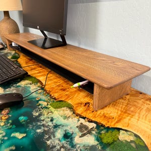 May include: A wooden monitor stand with a black monitor on top. The stand has a built-in cable management system and a small drawer. The stand is sitting on a desk with a blue and green epoxy resin surface.