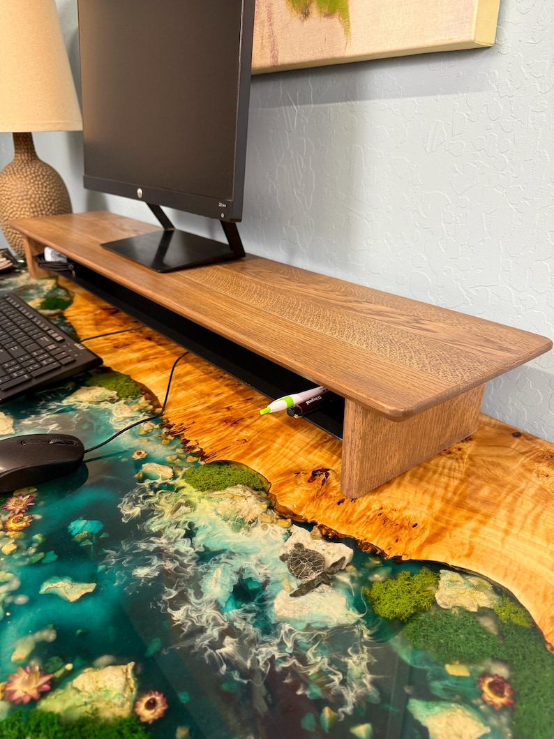 May include: A wooden monitor stand with a black monitor on top. The stand has a built-in cable management system and a small drawer. The stand is sitting on a desk with a blue and green epoxy resin surface.