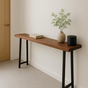 May include: A long, wooden console table with black metal legs. The table is decorated with a book and a vase with green plants.