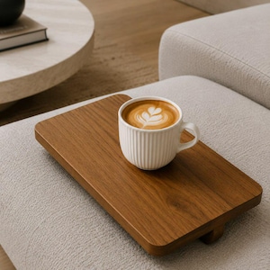 May include: A white ceramic coffee cup with a latte art design sits on a rectangular wooden tray. The tray is a warm brown color and rests on a light-colored upholstered surface. The cup has vertical ribbed detailing.