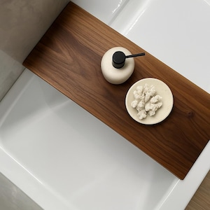 May include: A dark brown wooden bath tray with a cream-colored soap dispenser with a black pump and a small, round, cream-colored dish with white, coral-like objects. The tray is placed over a white bathtub.