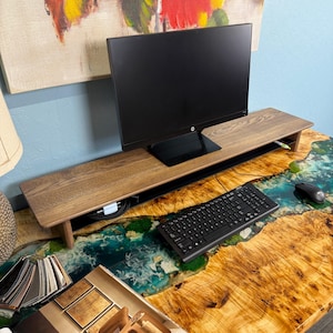 May include: A wooden monitor stand with a black computer monitor on top. The stand is on a desk with a resin river design and a keyboard and mouse.
