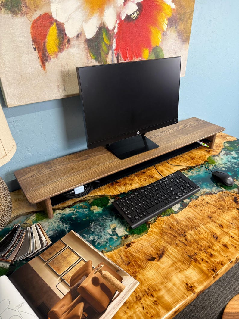 May include: A wooden monitor stand with a black computer monitor on top. The stand is on a desk with a resin river design and a keyboard and mouse.