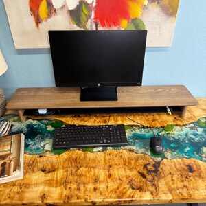May include: A wooden desk with a live edge epoxy river design. The desk has a black computer monitor, keyboard, and mouse on top. The desk is in front of a wall with a painting of flowers.