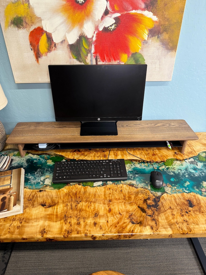 May include: A wooden desk with a live edge epoxy river design. The desk has a black computer monitor, keyboard, and mouse on top. The desk is in front of a wall with a painting of flowers.