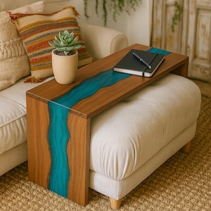 May include: A wooden side table with a teal river design. The rectangular table sits on a beige ottoman. A succulent in a beige pot, a black notebook, and a pen are on the table. The table is made of wood and epoxy resin.