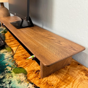 May include: A wooden monitor stand with a shelf for storage. The stand is made of light brown wood and has a natural finish. The stand is sitting on a desk with a blue and green epoxy resin surface.