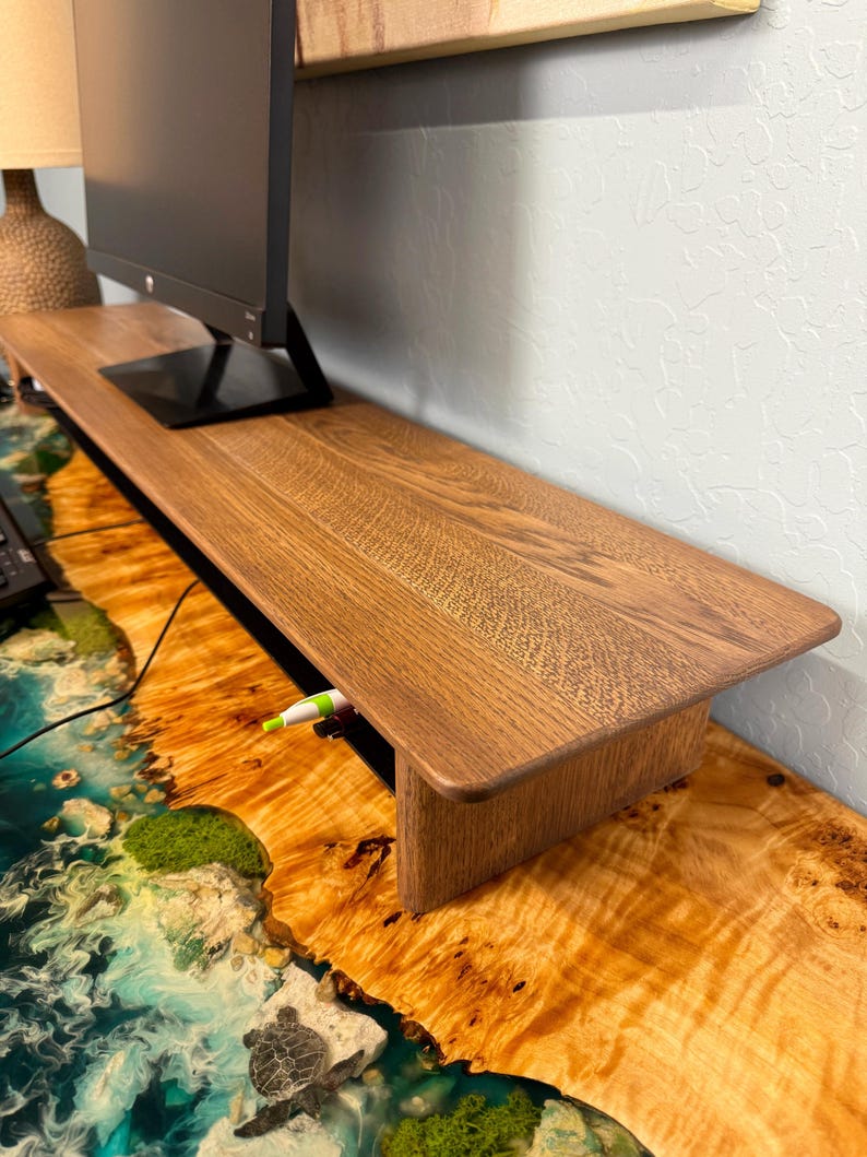 May include: A wooden monitor stand with a shelf for storage. The stand is made of light brown wood and has a natural finish. The stand is sitting on a desk with a blue and green epoxy resin surface.