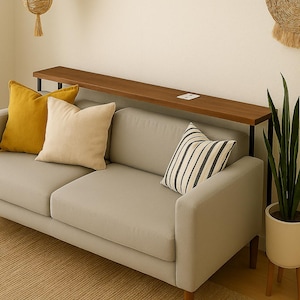 Puede incluir: Un sofá gris moderno con patas de madera, adornado con cojines amarillos, crema y a rayas. Una larga estantería de madera oscura se encuentra detrás del sofá. Una planta en maceta y un tapiz de pared tejido completan la decoración.