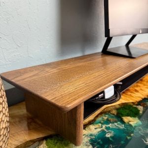 May include: A wooden monitor stand with a black wire management shelf. The stand is made of dark wood and has a smooth finish. The stand is sitting on a blue and green epoxy resin surface.