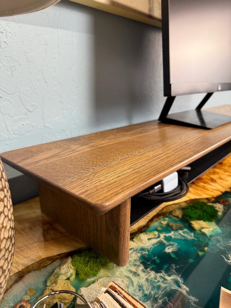 May include: A wooden monitor stand with a black wire management shelf. The stand is made of dark wood and has a smooth finish. The stand is sitting on a blue and green epoxy resin surface.