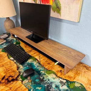 May include: A wooden monitor stand with a cable management system sits on a desk with a unique epoxy river design. The desk is made of wood with a natural grain pattern. The monitor stand is a light brown colour and has a black edge. The desk has a blue, green, and white epoxy river design.
