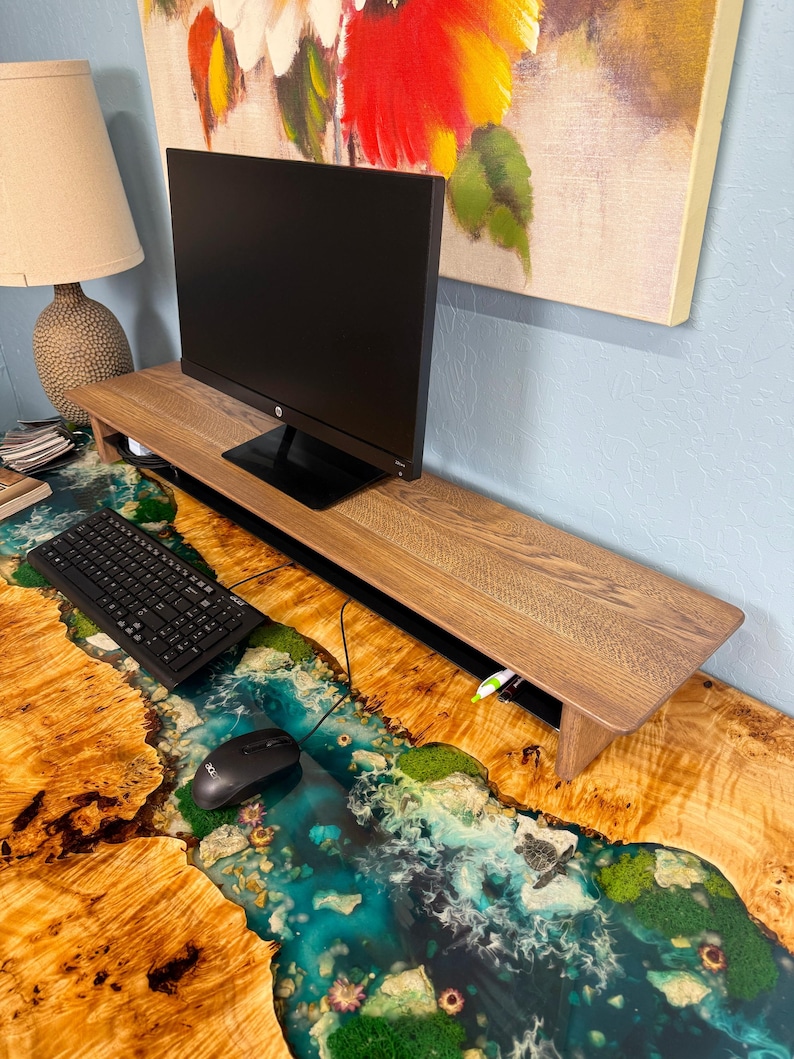 May include: A wooden monitor stand with a cable management system sits on a desk with a unique epoxy river design. The desk is made of wood with a natural grain pattern. The monitor stand is a light brown colour and has a black edge. The desk has a blue, green, and white epoxy river design.