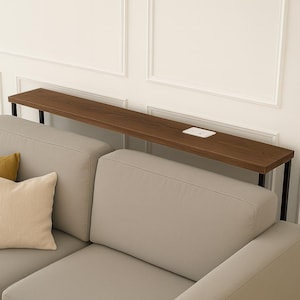 Puede incluir: Una consola moderna de madera marrón con marco de metal negro, colocada detrás de un sofá gris claro. La mesa tiene una toma de corriente integrada. La pared detrás de la mesa es blanca con molduras decorativas.