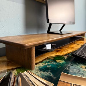 May include: A wooden monitor stand with a black metal shelf. The stand is sitting on a desk with a blue and green epoxy resin surface. There are also some fabric swatches on the desk.