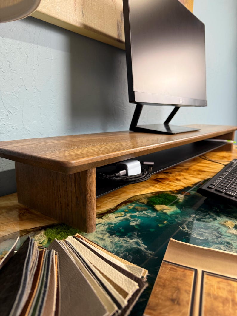 May include: A wooden monitor stand with a black metal shelf. The stand is sitting on a desk with a blue and green epoxy resin surface. There are also some fabric swatches on the desk.