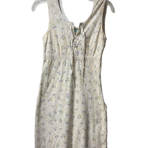 May include: A sleeveless, cream-colored dress with a floral pattern of blue and yellow flowers. The dress features a lace-up neckline, a fitted bodice, and a straight skirt. The hemline is trimmed with delicate lace.