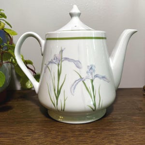 May include: White ceramic teapot with a lid, handle, and spout. The teapot features a green band around the top and is decorated with light purple iris flowers and green stems. The teapot sits on a wooden surface.