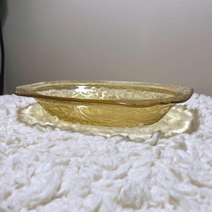 May include: A vintage, oval-shaped, amber-colored glass bowl with a decorative floral pattern. The bowl sits on a matching, slightly larger, oval-shaped plate. The glass has a textured surface and a scalloped edge.