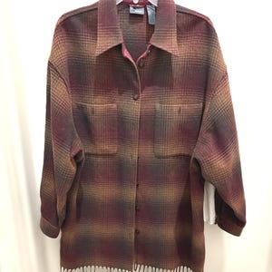 May include: A long-sleeved, button-down plaid shirt in shades of burgundy, brown, and tan. The shirt has two front pockets and a fringed hem. The collar and cuffs are visible. The shirt is hanging on a white hanger.