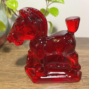 Puede incluir: Una vibrante figura de caballo de cristal rojo. El caballo está representado en posición sentada, con una melena y cola detalladas. La figura está sobre una base rectangular. El cristal tiene un acabado brillante, reflejando la luz.