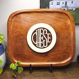May include: A rectangular wooden serving tray with rounded edges. A white circular coaster in the center features the word "CHEESE" in a brown, stylized font. The tray is made of a reddish-brown wood.
