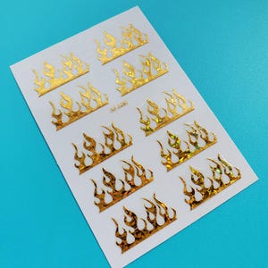 May include: A sheet of gold flame nail decals with a holographic finish. There are 10 flame decals on the sheet. The sheet is white and has the text "NF-328T" printed on it.