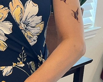 Puede incluir: Dos tatuajes temporales de mariposas aplicados en la parte superior del brazo. La mariposa superior presenta alas naranjas y amarillas, mientras que la inferior tiene tonos morados y azules. La piel está junto a un vestido azul marino con estampado floral amarillo.