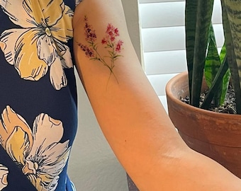 May include: A navy blue dress featuring a large-scale yellow and white floral pattern. On the upper arm, there is a delicate, colorful tattoo depicting two sprigs of small pink and purple wildflowers with thin green stems and leaves.