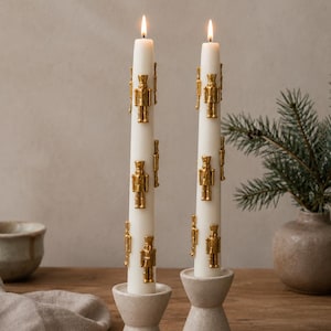 May include: Two white taper candles, each lit with a small flame, featuring multiple small, gold-colored nutcracker figures attached to the exterior. The candles are held upright in two matching, light-colored, textured, hourglass-shaped candle holders.